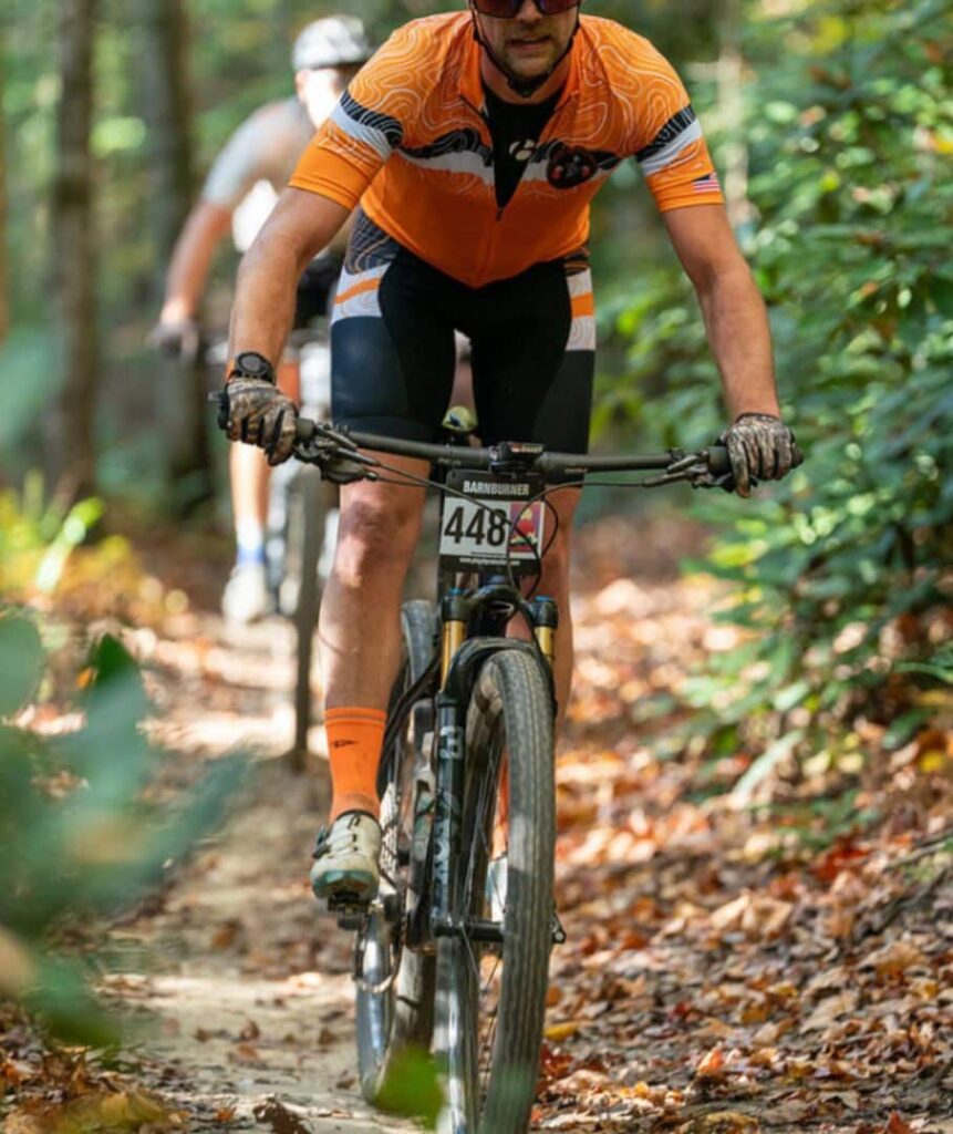 Cyclist in orange and black racing gear riding gravel or hybrid bike on paved downhill trail with wooden rail fence visible, navigating technical terrain during race.