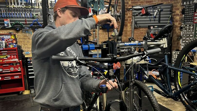 Professional bicycle mechanic performing detailed repair work on bike at workbench in Skinny Wheels Pedals & Pints shop, with tools and repair equipment visible.